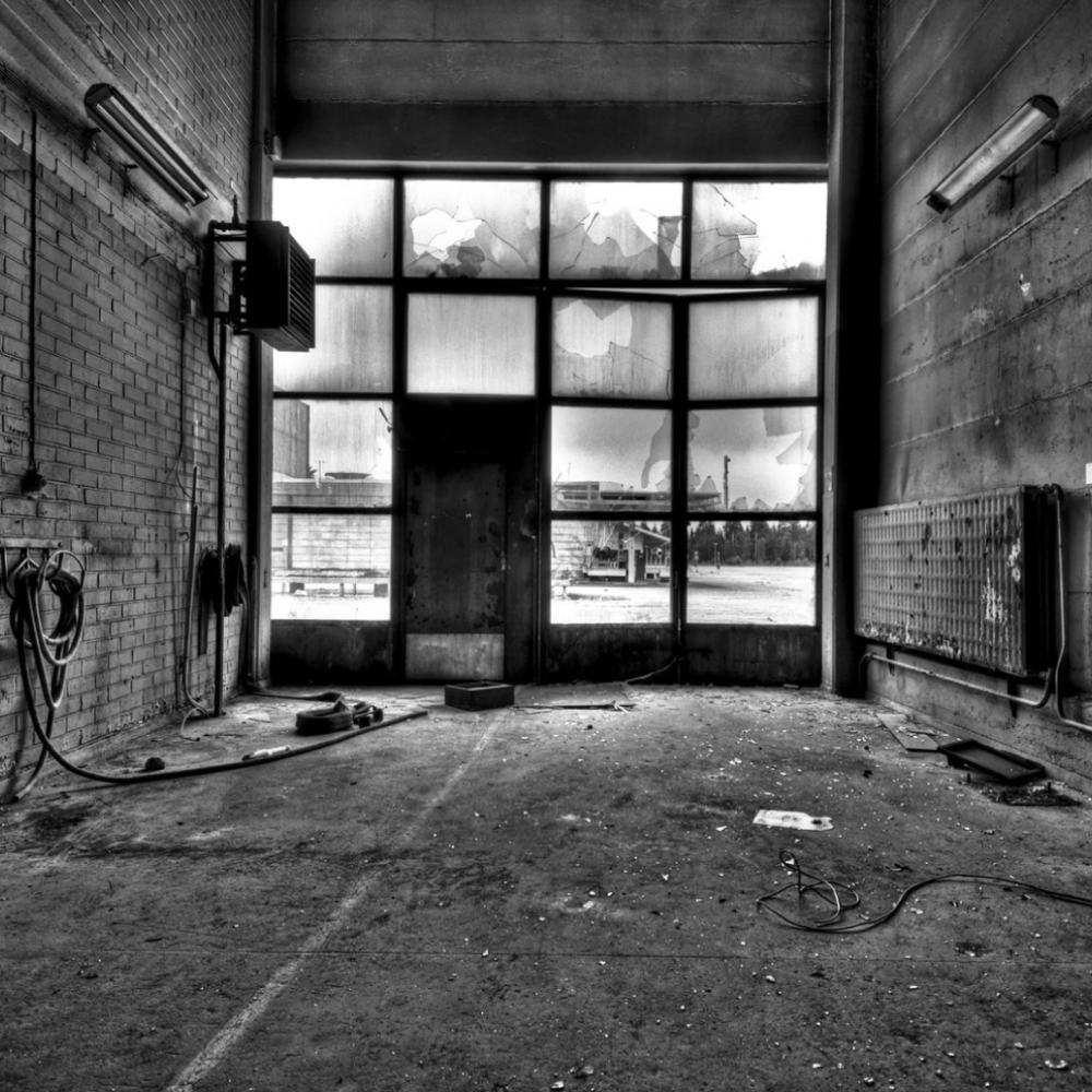 Svartvit bild av ett tomt, dammigt lager med krossade fönster, synliga tegelväggar och spritt skräp.