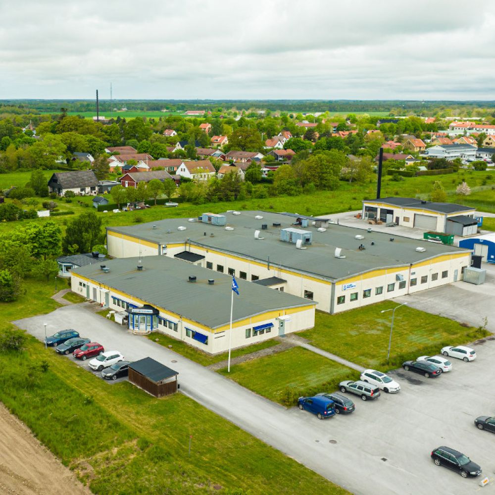 En flygvy över en stor industribyggnad med flera parkeringsplatser omgivna av gröna fält och bostadsområden.
